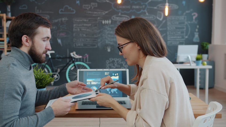Two colleagues discussing a project at a desk. business team workflow diagram, automation flowchart on screen, enterprise SaaS integration