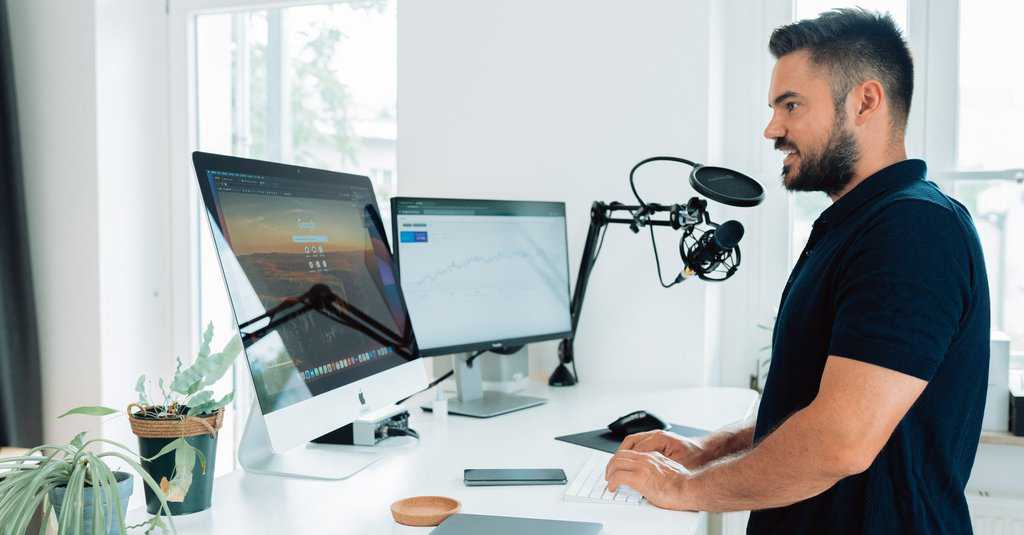Man at work with podcast microphone in front of a desk and two screens with splitscreen computer and laptop on the desk in a social media agency. Working on a computer.