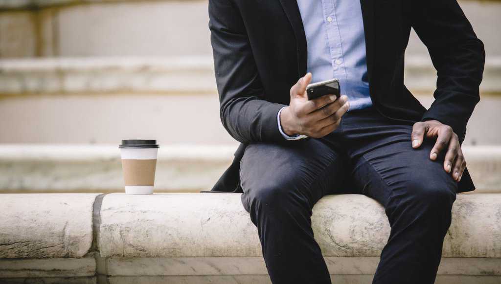 Crop businessman using smartphone while resting on bench with takeaway coffee