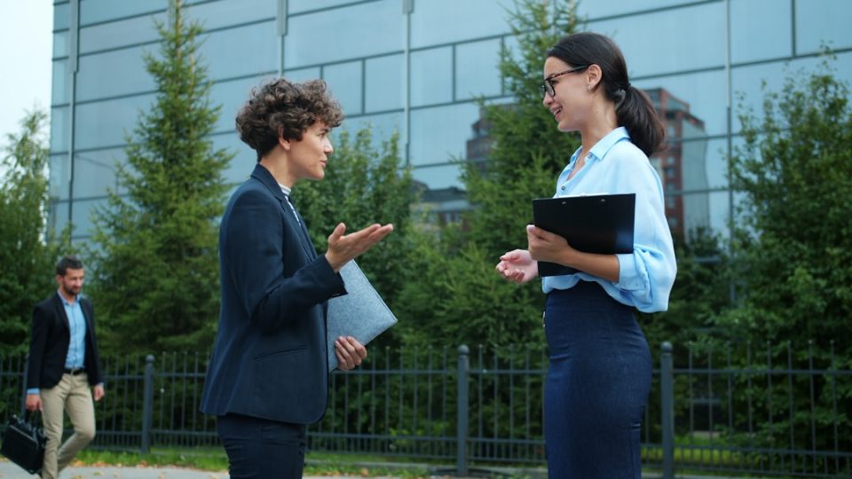 Two businesswomen talking outside modern office building. professional networking handshake, business meeting exchange card, corporate environment